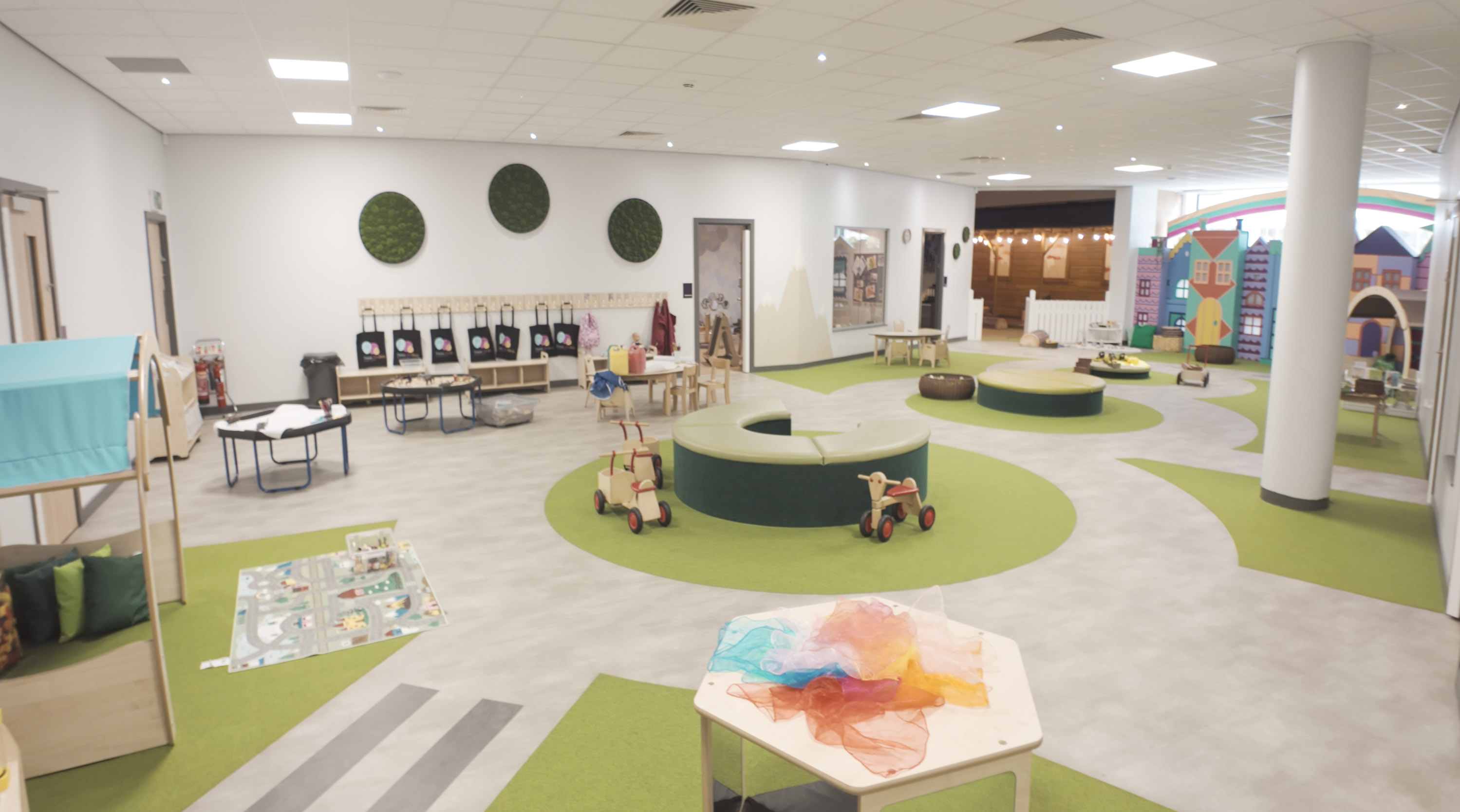 Large indoor garden-themed movement space with ride-on toys and a village mural